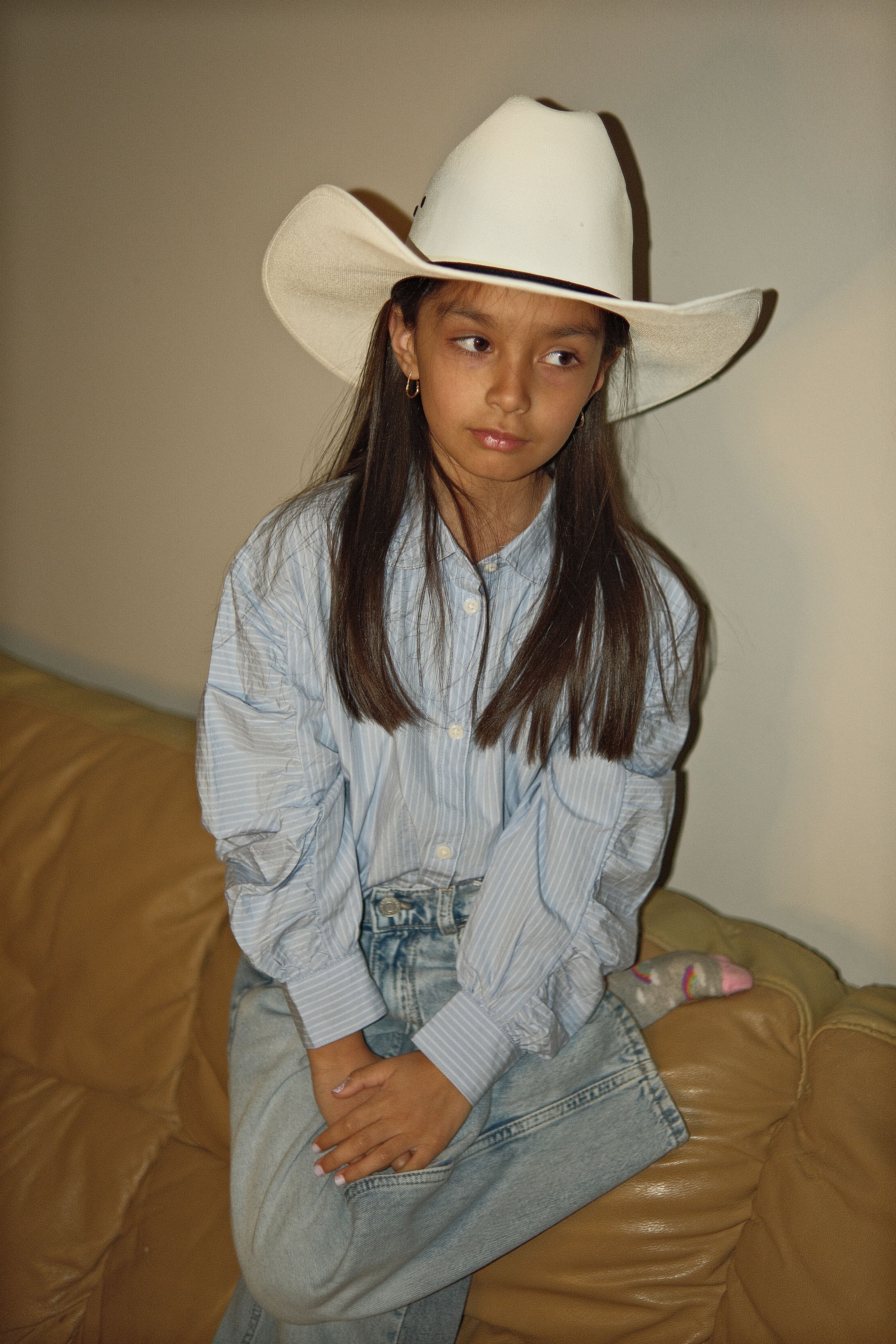 Young girl Solomika seated thoughtfully on a couch, wearing cowboy hat and striped shirt, indoor flash photo.