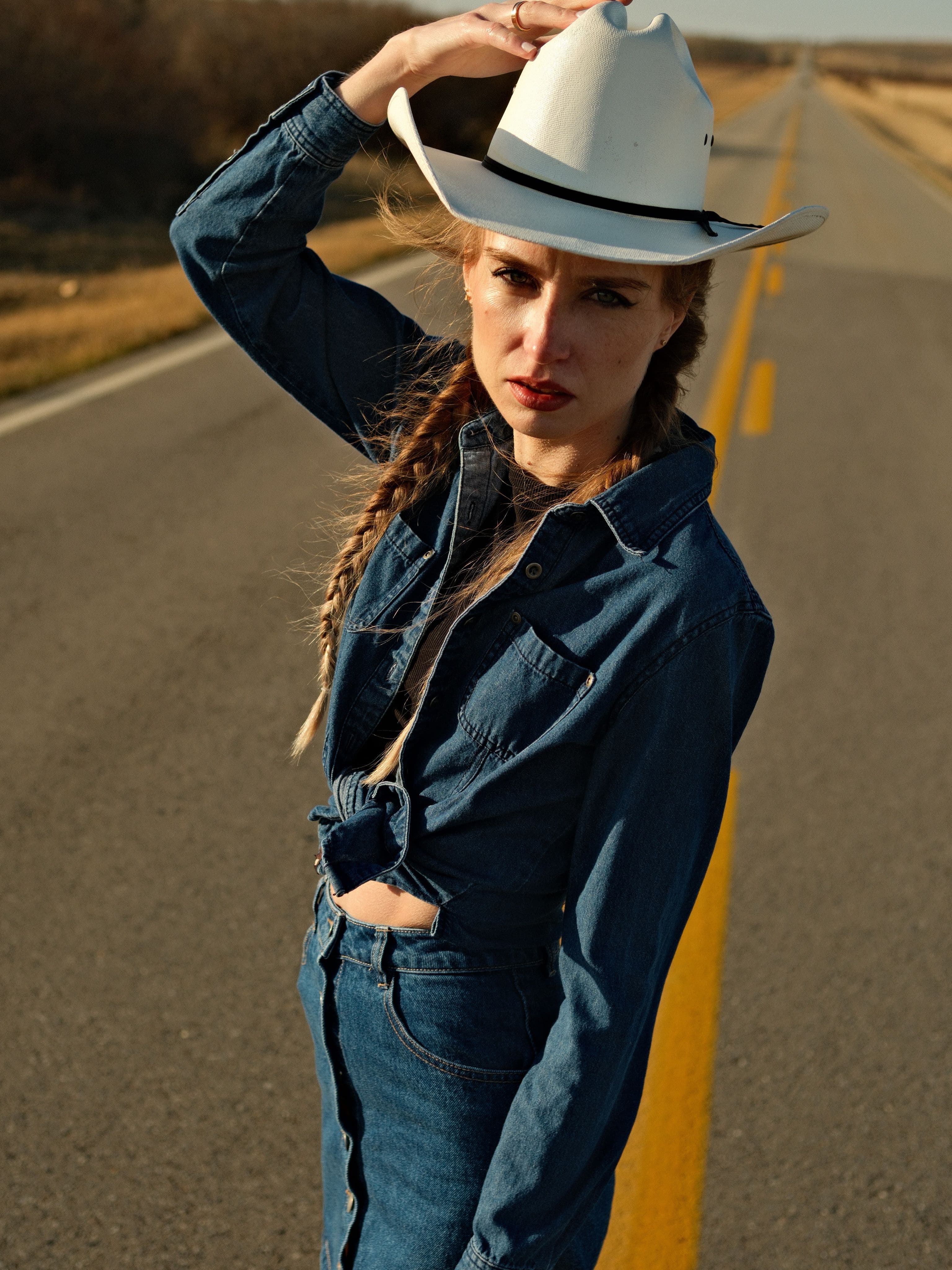 Close-up of model Mariana adjusting her white cowboy hat, wearing denim-on-denim outfit on rural highway on the outskirts of Cochrane,AB.